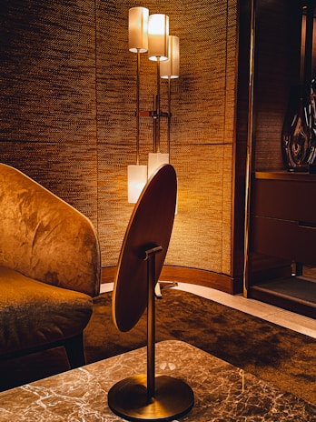 A warmly lit interior features a modern floor lamp with multiple cylindrical shades, casting a soft light against a textured wall. The room includes a plush armchair with rich fabric and a sleek wooden table with a marble top. The overall design aesthetics are sophisticated and cozy.