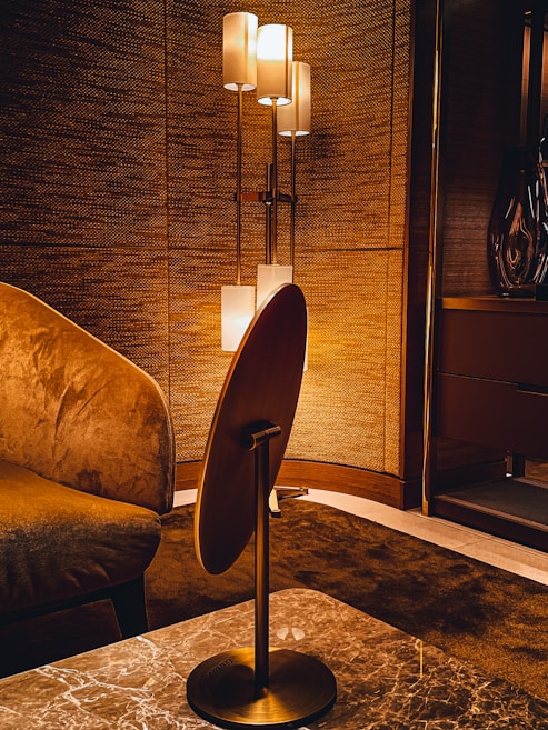 A warmly lit interior features a modern floor lamp with multiple cylindrical shades, casting a soft light against a textured wall. The room includes a plush armchair with rich fabric and a sleek wooden table with a marble top. The overall design aesthetics are sophisticated and cozy.