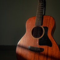 A wooden acoustic guitar stands against a dark green background, with light casting a soft shadow across its body. The guitar has a warm, rich brown color and features a prominent black pickguard. The strings and tuning pegs are clearly visible, accentuating the instrument's detailed craftsmanship.