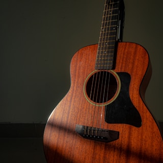 A wooden acoustic guitar stands against a dark green background, with light casting a soft shadow across its body. The guitar has a warm, rich brown color and features a prominent black pickguard. The strings and tuning pegs are clearly visible, accentuating the instrument's detailed craftsmanship.