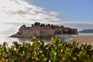 A picturesque island connected to the mainland by a narrow isthmus. The island is covered with buildings featuring red-tiled roofs, likely a mixture of historic stone structures surrounded by lush greenery. In the foreground, there is a sandy beach with a few people walking and enjoying the scenery. The calm sea reflects the partly cloudy sky, creating a serene atmosphere.