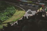 A rally car speeds along a curving track surrounded by lush greenery. Spectators fill the stands watching the race with anticipation. Safety personnel in orange outfits are visible near the track barriers.