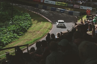 A rally car speeds along a curving track surrounded by lush greenery. Spectators fill the stands watching the race with anticipation. Safety personnel in orange outfits are visible near the track barriers.