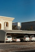 A row of white cars is parked under a metal canopy next to a beige building with decorative columns and a small balcony. The sky is clear and blue, and there are palm trees visible in the background.