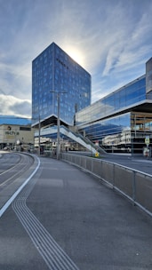 A modern cityscape featuring a tall, reflective skyscraper with sunlight shining from behind. The foreground shows a tram line curving alongside a pedestrian path. Glass facades and an elevated pedestrian walkway connect buildings, reflecting the cloudy sky.