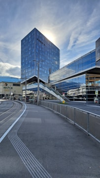 A modern cityscape featuring a tall, reflective skyscraper with sunlight shining from behind. The foreground shows a tram line curving alongside a pedestrian path. Glass facades and an elevated pedestrian walkway connect buildings, reflecting the cloudy sky.
