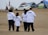 A group of four people wearing white T-shirts with #NODAPL printed on the back walk on a sandy area. There are tents and a car in the background, possibly indicating a campsite or protest site. The sky is overcast, and the setting has a modest, makeshift appearance.