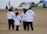 A group of four people wearing white T-shirts with #NODAPL printed on the back walk on a sandy area. There are tents and a car in the background, possibly indicating a campsite or protest site. The sky is overcast, and the setting has a modest, makeshift appearance.
