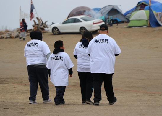 A group of four people wearing white T-shirts with #NODAPL printed on the back walk on a sandy area. There are tents and a car in the background, possibly indicating a campsite or protest site. The sky is overcast, and the setting has a modest, makeshift appearance.
