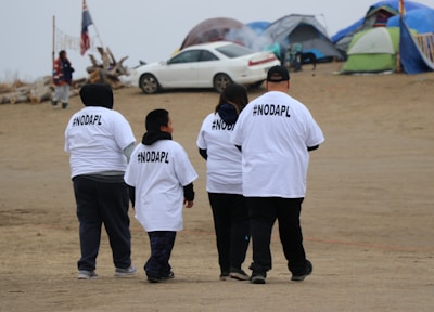 A group of four people wearing white T-shirts with #NODAPL printed on the back walk on a sandy area. There are tents and a car in the background, possibly indicating a campsite or protest site. The sky is overcast, and the setting has a modest, makeshift appearance.