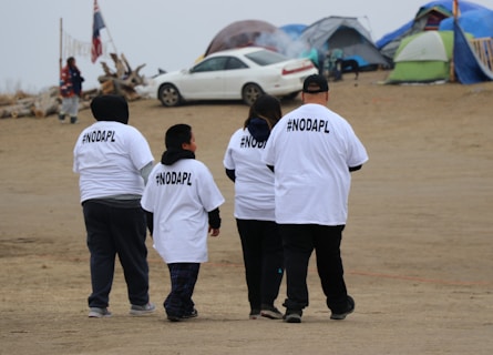 A group of four people wearing white T-shirts with #NODAPL printed on the back walk on a sandy area. There are tents and a car in the background, possibly indicating a campsite or protest site. The sky is overcast, and the setting has a modest, makeshift appearance.