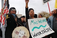 A group of people are participating in a protest or rally. One person is holding a sign that reads 'Mni Wiconi, Water is Life.' Another person is holding a drum with a painted buffalo. The atmosphere suggests a sense of solidarity and activism. There are multiple flags, including a U.S. flag, visible in the scene.