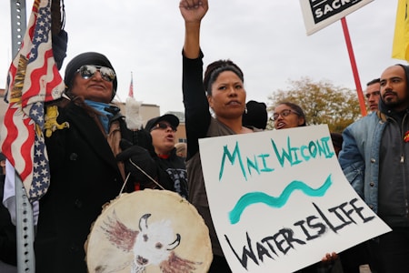 A group of people are participating in a protest or rally. One person is holding a sign that reads 'Mni Wiconi, Water is Life.' Another person is holding a drum with a painted buffalo. The atmosphere suggests a sense of solidarity and activism. There are multiple flags, including a U.S. flag, visible in the scene.