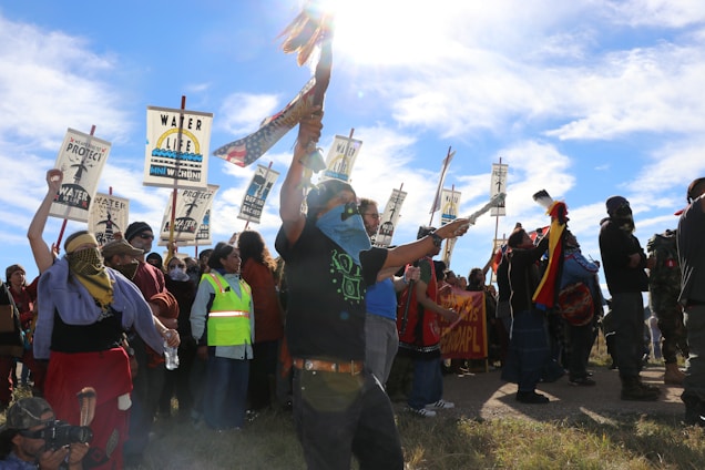 A group of people is gathered in a protest or demonstration, holding signs that advocate for water protection and environmental rights. Various individuals are visible, some wearing traditional clothing and others in casual attire. The atmosphere seems energetic and purposeful, enhanced by the bright, sunny day and a clear blue sky.