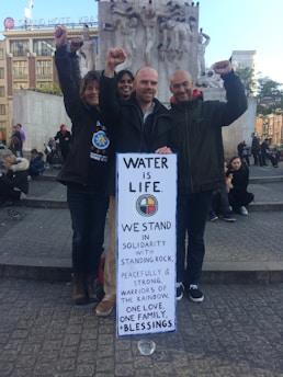 Three people are standing and smiling while holding up a sign that reads 'Water is Life. We stand in solidarity with Standing Rock. Peacefully and strong. Warriors of the rainbow. One love. One family. Blessings.' They are raising their fists in a gesture of protest. In the background, there is a monument and people sitting around in a public square.