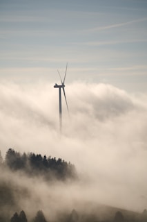 A wind turbine rises above a thick layer of fog, with its blades silhouetted against the sky. Below, a forested area is partially visible through the mist, adding a sense of tranquility and mystery to the scene.
