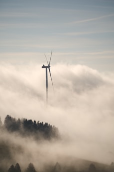 A wind turbine rises above a thick layer of fog, with its blades silhouetted against the sky. Below, a forested area is partially visible through the mist, adding a sense of tranquility and mystery to the scene.