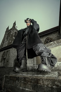 A person dressed in dark, futuristic clothing stands in front of a historical stone building with Gothic architecture, wearing large boots and sunglasses. The overcast sky creates a dramatic backdrop.