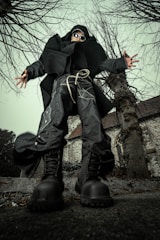 A person dressed in avant-garde, futuristic clothing stands in a dynamic pose from a low-angle perspective. The individual is wearing oversized black boots, baggy pants with intricate patterns, a large black coat, and sunglasses. They are surrounded by leafless trees and an old, stone building with a steep roof and an arched window in the background.