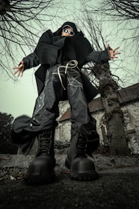 A person dressed in avant-garde, futuristic clothing stands in a dynamic pose from a low-angle perspective. The individual is wearing oversized black boots, baggy pants with intricate patterns, a large black coat, and sunglasses. They are surrounded by leafless trees and an old, stone building with a steep roof and an arched window in the background.
