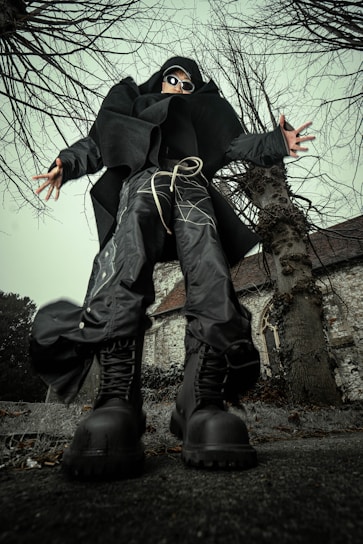 A person dressed in avant-garde, futuristic clothing stands in a dynamic pose from a low-angle perspective. The individual is wearing oversized black boots, baggy pants with intricate patterns, a large black coat, and sunglasses. They are surrounded by leafless trees and an old, stone building with a steep roof and an arched window in the background.
