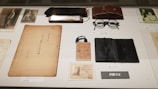 A collection of vintage items is displayed on a surface. Items include a harmonica, a pair of round spectacles, a worn book or document, an old identification card, photographs, and a leather case. The items are meticulously arranged and labeled, suggesting a historical or personal significance.