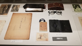 A collection of vintage items is displayed on a surface. Items include a harmonica, a pair of round spectacles, a worn book or document, an old identification card, photographs, and a leather case. The items are meticulously arranged and labeled, suggesting a historical or personal significance.