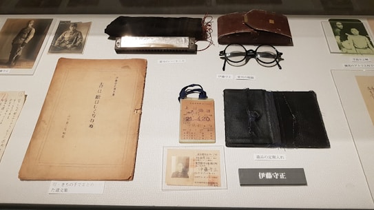 A collection of vintage items is displayed on a surface. Items include a harmonica, a pair of round spectacles, a worn book or document, an old identification card, photographs, and a leather case. The items are meticulously arranged and labeled, suggesting a historical or personal significance.