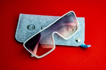 A pair of oversized sunglasses with white frames and tinted lenses rests on top of a light gray felt sunglasses case. The background is solid red, providing contrast to the case and the shades.