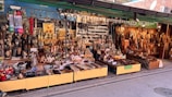 A vibrant street market stall displays a wide assortment of handcrafted goods. There are carved wooden items, ornate figurines, decorative objects, and kitchen utensils neatly arranged on tables and shelves. The setup is colorful, with various textures and materials, such as metal, wood, and ceramics. Signs with Korean language text are visible, indicating pricing or descriptions of the items.