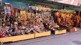 A vibrant street market stall displays a wide assortment of handcrafted goods. There are carved wooden items, ornate figurines, decorative objects, and kitchen utensils neatly arranged on tables and shelves. The setup is colorful, with various textures and materials, such as metal, wood, and ceramics. Signs with Korean language text are visible, indicating pricing or descriptions of the items.
