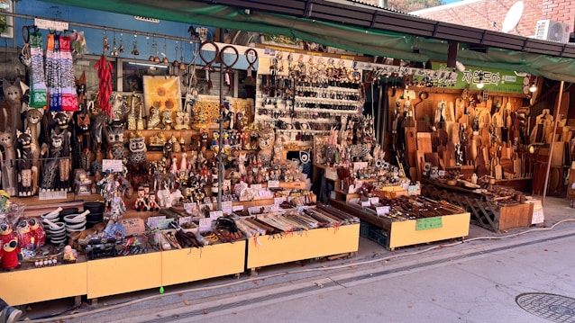 A vibrant street market stall displays a wide assortment of handcrafted goods. There are carved wooden items, ornate figurines, decorative objects, and kitchen utensils neatly arranged on tables and shelves. The setup is colorful, with various textures and materials, such as metal, wood, and ceramics. Signs with Korean language text are visible, indicating pricing or descriptions of the items.