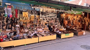 A vibrant street market stall displays a wide assortment of handcrafted goods. There are carved wooden items, ornate figurines, decorative objects, and kitchen utensils neatly arranged on tables and shelves. The setup is colorful, with various textures and materials, such as metal, wood, and ceramics. Signs with Korean language text are visible, indicating pricing or descriptions of the items.
