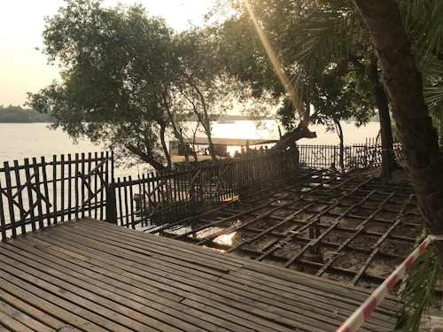 A serene riverside scene featuring a wooden deck and railing. Lush green trees partially shade the area, with sunlight filtering through, casting reflections on the water's surface. The river flows gently in the background.