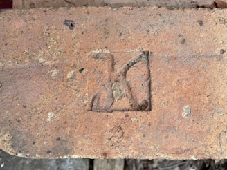 A weathered brick with a stamped mark that resembles a letter or symbol in a recessed rectangular area. The brick is earthy in color, showing signs of age with small patches of discoloration and rough texture.