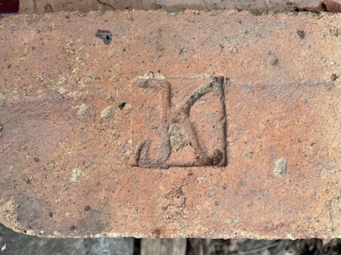 A weathered brick with a stamped mark that resembles a letter or symbol in a recessed rectangular area. The brick is earthy in color, showing signs of age with small patches of discoloration and rough texture.