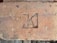 A weathered brick with a stamped mark that resembles a letter or symbol in a recessed rectangular area. The brick is earthy in color, showing signs of age with small patches of discoloration and rough texture.