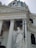 A grand architectural structure featuring a classical building with tall columns and intricate moldings. A prominent statue of an angel holding a large cross stands in front, with a large dome and smaller statues on top. The facade includes Latin inscriptions.