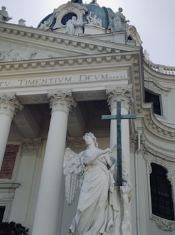 A grand architectural structure featuring a classical building with tall columns and intricate moldings. A prominent statue of an angel holding a large cross stands in front, with a large dome and smaller statues on top. The facade includes Latin inscriptions.