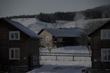 A snowy landscape features several wooden cabins, with smoke rising from the chimneys, indicating the warmth inside despite the chilly surroundings. In the background, a ski slope can be seen with traces of skier activity. The scene is framed by leafless trees and a forest in the distance.