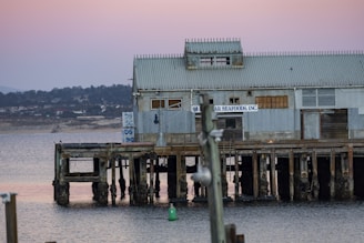 An old, weathered structure stands elevated above the water on wooden stilts. The building has a corrugated metal roof and walls, with a sign indicating 'Seafoods, Inc.' The backdrop reveals a hazy view of distant hills and a peaceful water surface under a soft, pastel-colored sky.