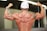 A muscular person is flexing their back and arm muscles, wearing a white cap and displaying prominent tattoos on both arms. The skin appears shiny as if from sweat, indicating physical exertion. A watch is worn on the left wrist.