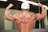 A muscular person is flexing their back and arm muscles, wearing a white cap and displaying prominent tattoos on both arms. The skin appears shiny as if from sweat, indicating physical exertion. A watch is worn on the left wrist.
