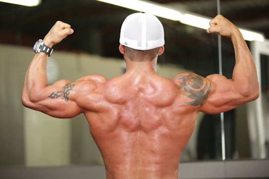 A muscular person is flexing their back and arm muscles, wearing a white cap and displaying prominent tattoos on both arms. The skin appears shiny as if from sweat, indicating physical exertion. A watch is worn on the left wrist.