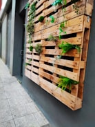 A vertical wooden pallet wall planter adorned with various small green plants and moss, installed on a smooth, gray concrete exterior wall along a sidewalk. The walkway is made of light gray tiles.