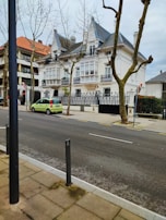 A residential street with a large, elegant house featuring white walls and intricate black wrought iron balconies. Tall trees with barren branches line the sidewalk, and a bright green car is parked on the street. The house possesses steep, slate-tiled roofs with dormer windows. The scene is urban, with a clear and clean street in the foreground.