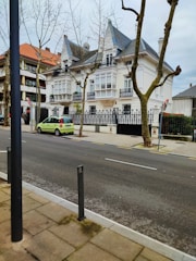 A residential street with a large, elegant house featuring white walls and intricate black wrought iron balconies. Tall trees with barren branches line the sidewalk, and a bright green car is parked on the street. The house possesses steep, slate-tiled roofs with dormer windows. The scene is urban, with a clear and clean street in the foreground.
