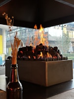 A modern fireplace with a rectangular metal frame contains evenly stacked dark stones, with bright yellow and orange flames rising above. Beyond the fireplace, there's a clear view of an urban setting through large glass windows, showcasing some outdoor greenery and architectural elements. In the foreground, a dark bottle holds dry ornamental grasses.