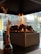 A modern fireplace with a rectangular metal frame contains evenly stacked dark stones, with bright yellow and orange flames rising above. Beyond the fireplace, there's a clear view of an urban setting through large glass windows, showcasing some outdoor greenery and architectural elements. In the foreground, a dark bottle holds dry ornamental grasses.
