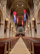 A grand, expansive church interior features rows of wooden pews flanking a long central aisle leading towards the altar. Tall, arched ceilings are adorned with hanging banners in blue, red, and gold hues. Large, ornate chandeliers illuminate the space with a warm glow, while intricate stone architecture displays elegant columns and details.